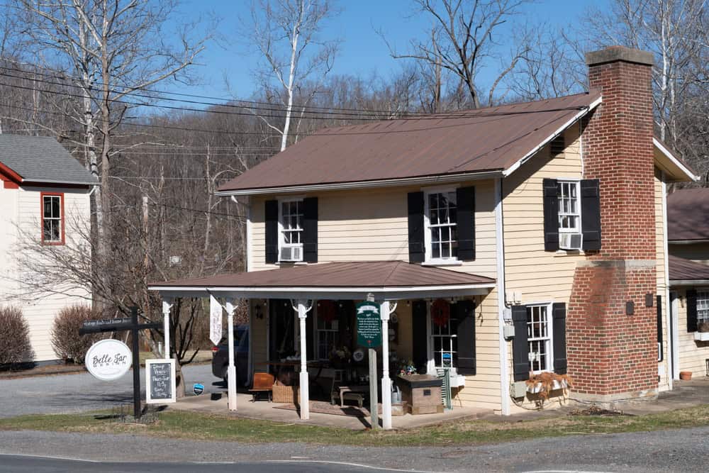 Belle Jin Gift Shop: A charming cream-colored historic house with black shutters, brick chimney, and welcoming front porch converted into a cozy rural store, nestled among winter-bare trees.