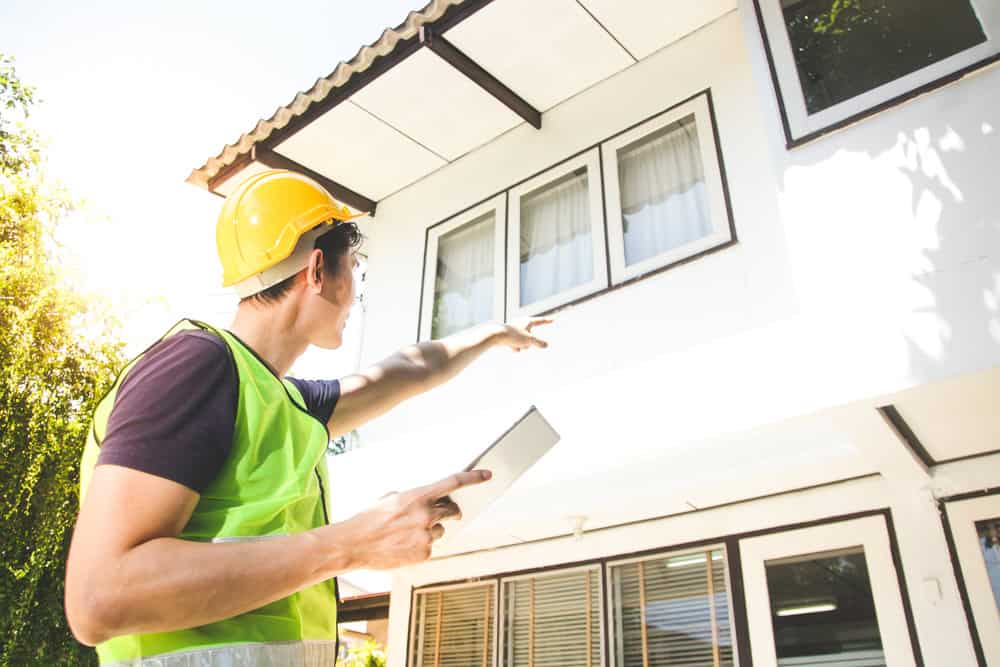 A construction worker inspects a building facade, using a tablet to take notes.