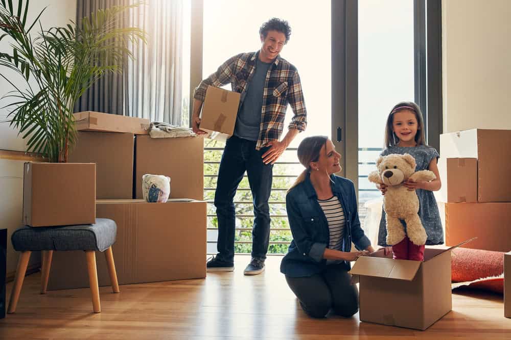 A family moves into their new home, surrounded by boxes and unpacking cherished items like a teddy bear.