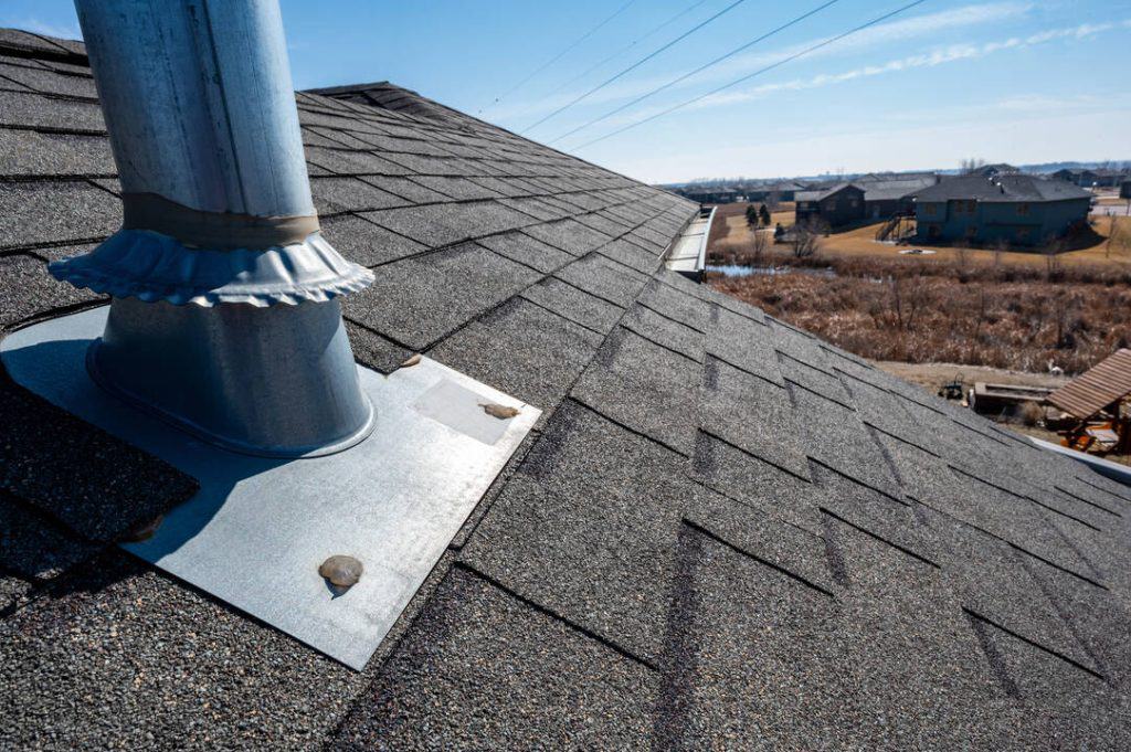 Close-up of a vent pipe flashing on a shingled suburban rooftop under a clear blue sky.