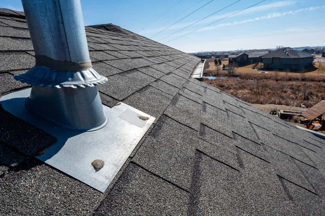 Close-up of a vent pipe flashing on a shingled suburban rooftop under a clear blue sky.