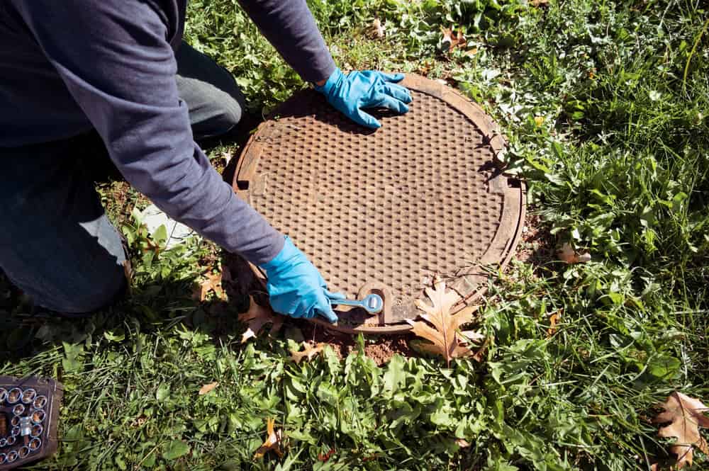 Worker with blue gloves opens a manhole cover in a grassy area for underground utility maintenance.
