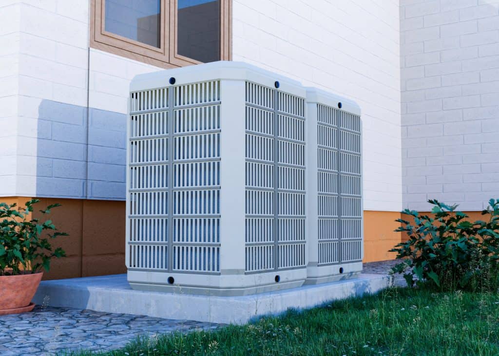 Two large central air conditioning units sit outside a house on a concrete pad, surrounded by garden plants and green grass.
