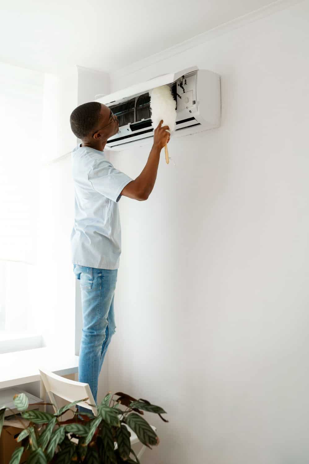 A man stands on a chair cleaning an air conditioner unit with a duster in a bright, modern room.