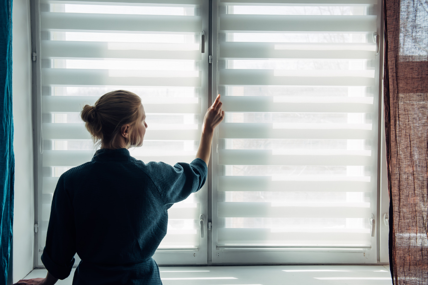 A woman in a dark robe adjusts striped window blinds as soft morning light fills the room.