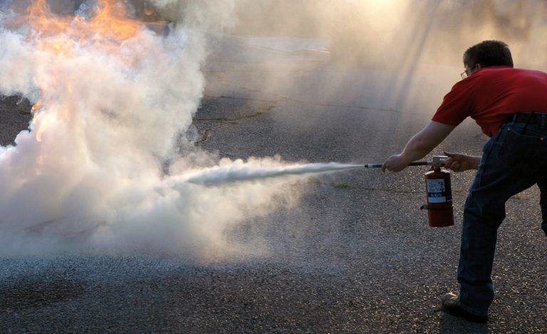 A man in a red shirt uses a fire extinguisher to battle flames on an asphalt surface, surrounded by thick smoke.