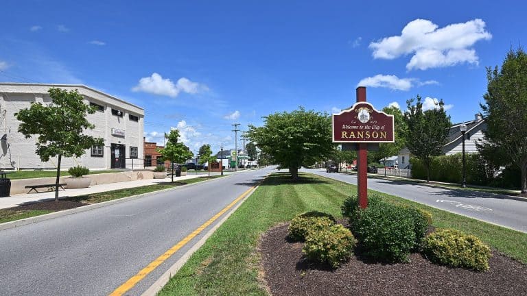 A sunny day welcomes visitors to Ranson, West Virginia, with its charming main street and a red city sign set against a vibrant blue sky.