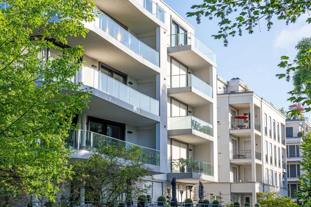 Modern apartment buildings with spacious glass balconies nestled among lush green trees on a sunny day.