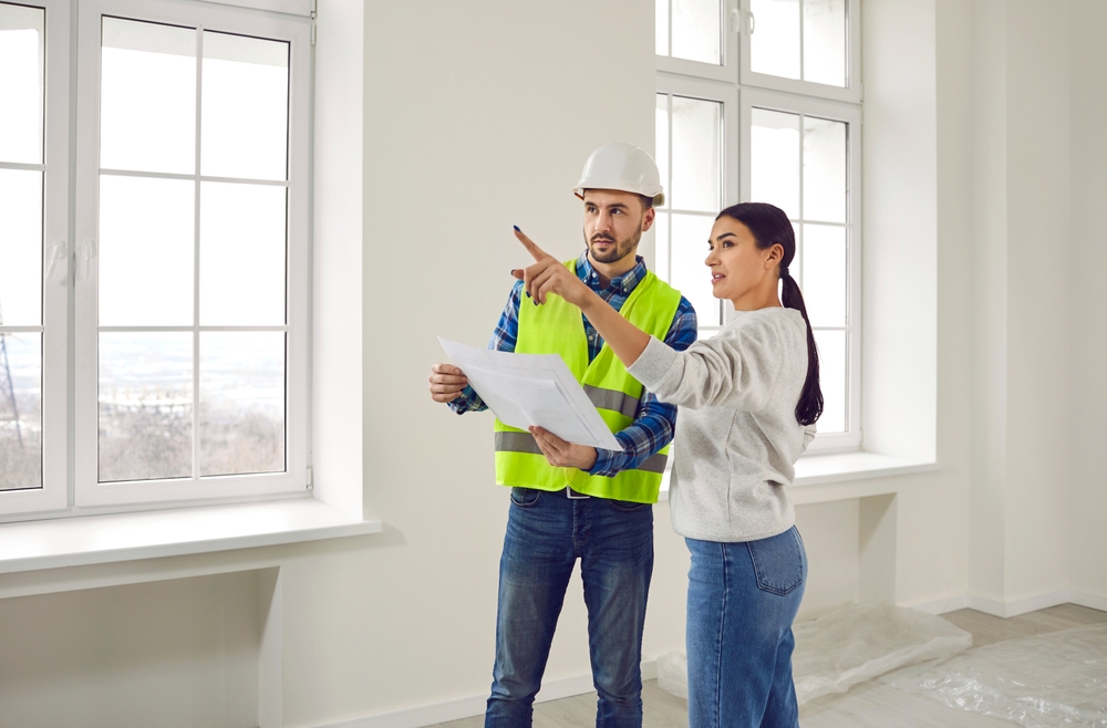 A woman discusses renovation plans with a construction worker inside a bright, modern room, pointing out details as he reviews blueprints.