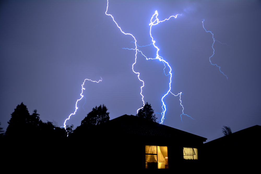 Lightning streaks dramatically across the night sky above a silhouetted house, its warm windows glowing against the storm.