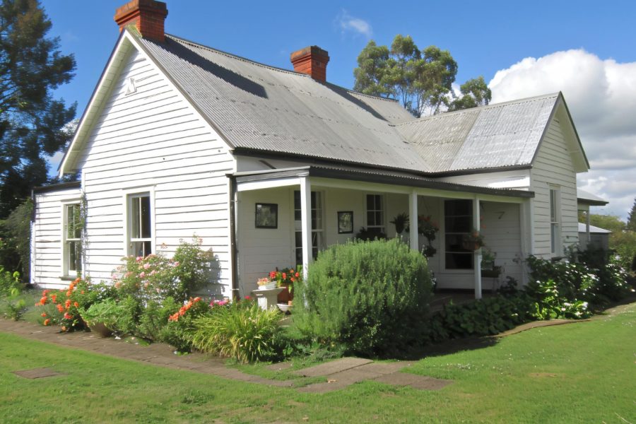 WV Home Charming white country cottage with a tin roof, surrounded by lush gardens and vibrant flowers on a sunny day.
