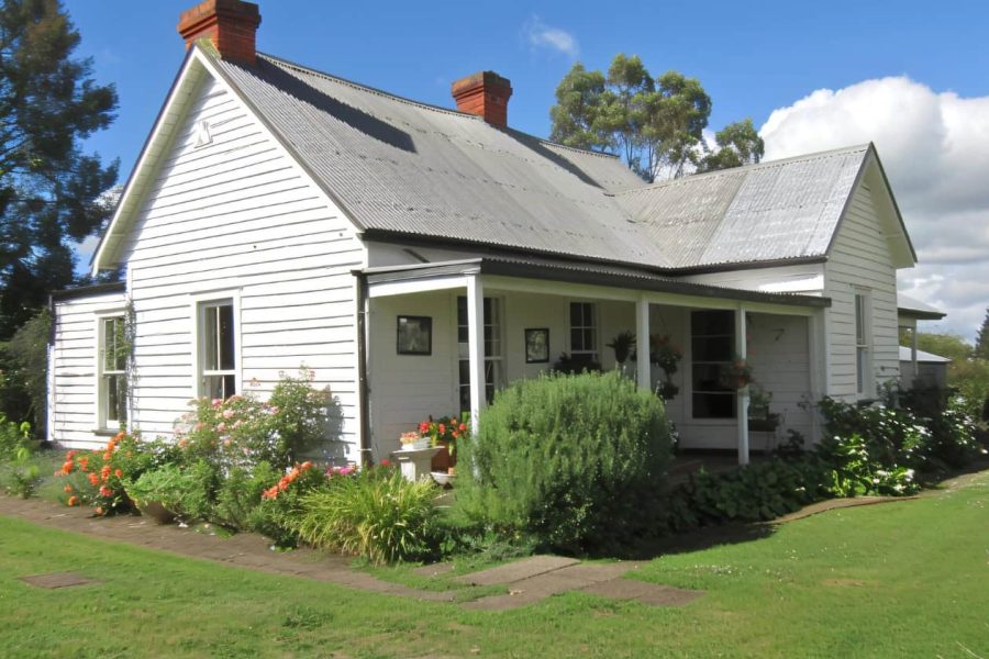 Charming white country cottage with a tin roof, surrounded by lush gardens and vibrant flowers on a sunny day.